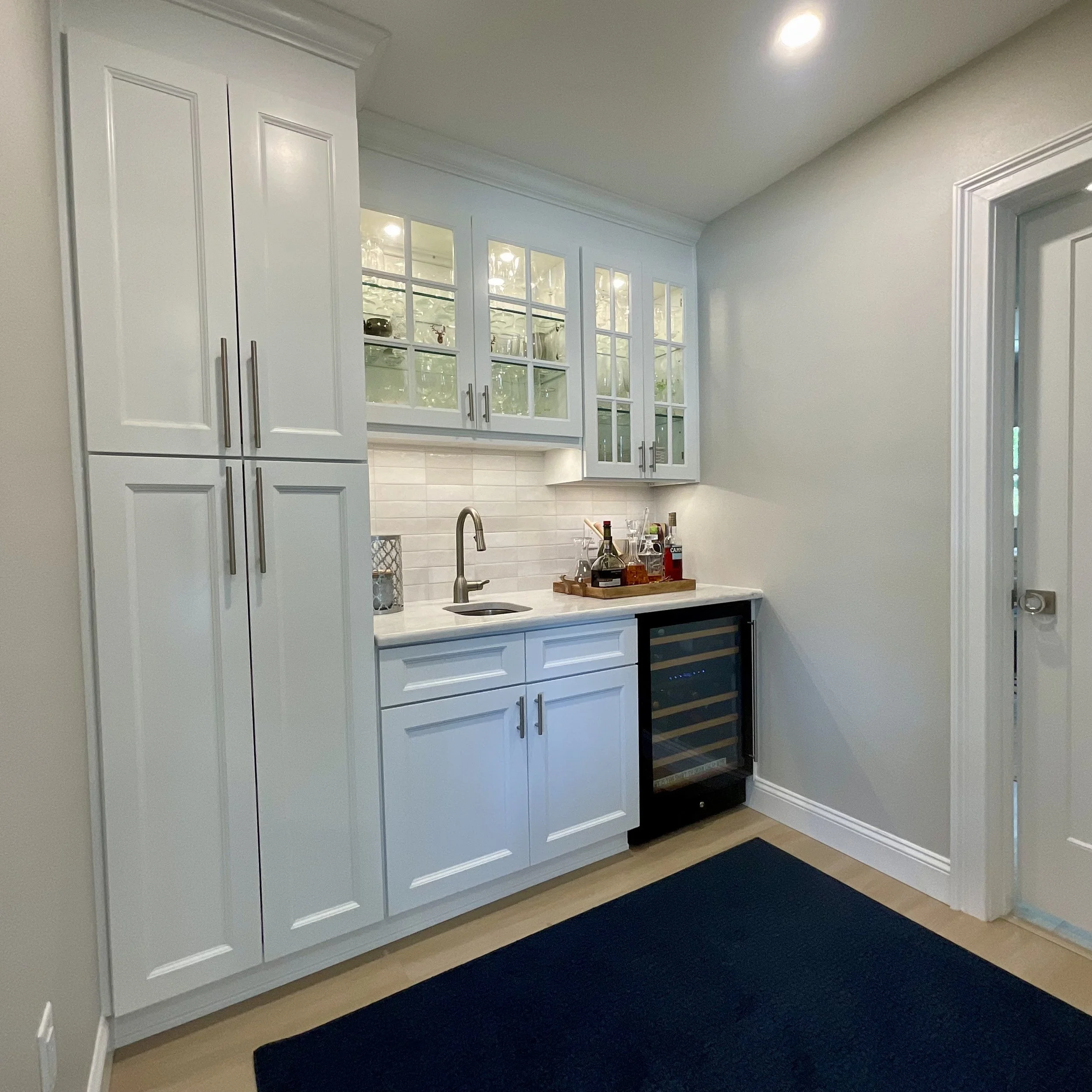 Modern wet bar with tall storage cabinet, small sink cabinet, under-counter beverage fridge, glass-front upper cabinets, and tile backsplash next to living room.