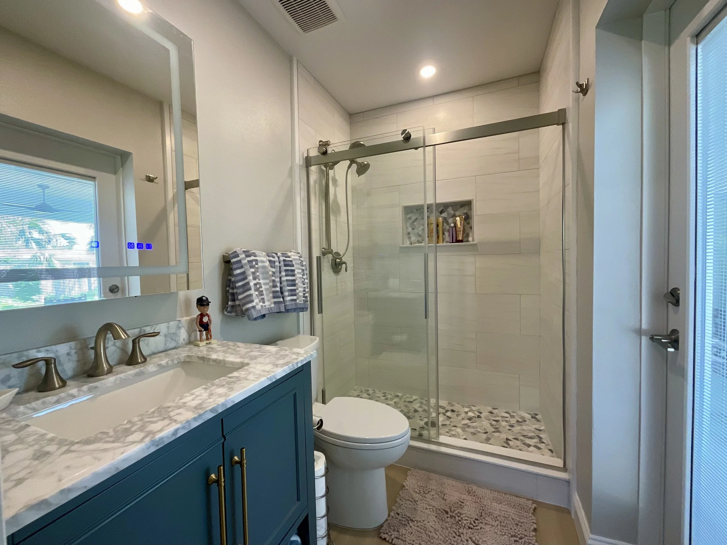 Updated pool bathroom with modern glass shower, large mirrored medicine cabinet, and full glass door opening to lanai, allowing natural light and easy outdoor access.
