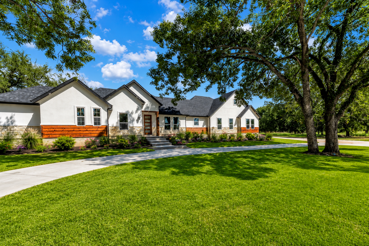 Newly constructed house with a white exterior, stone accents, and multiple windows surrounded by trees, with shadows and sandy yard in front under a blue sky.