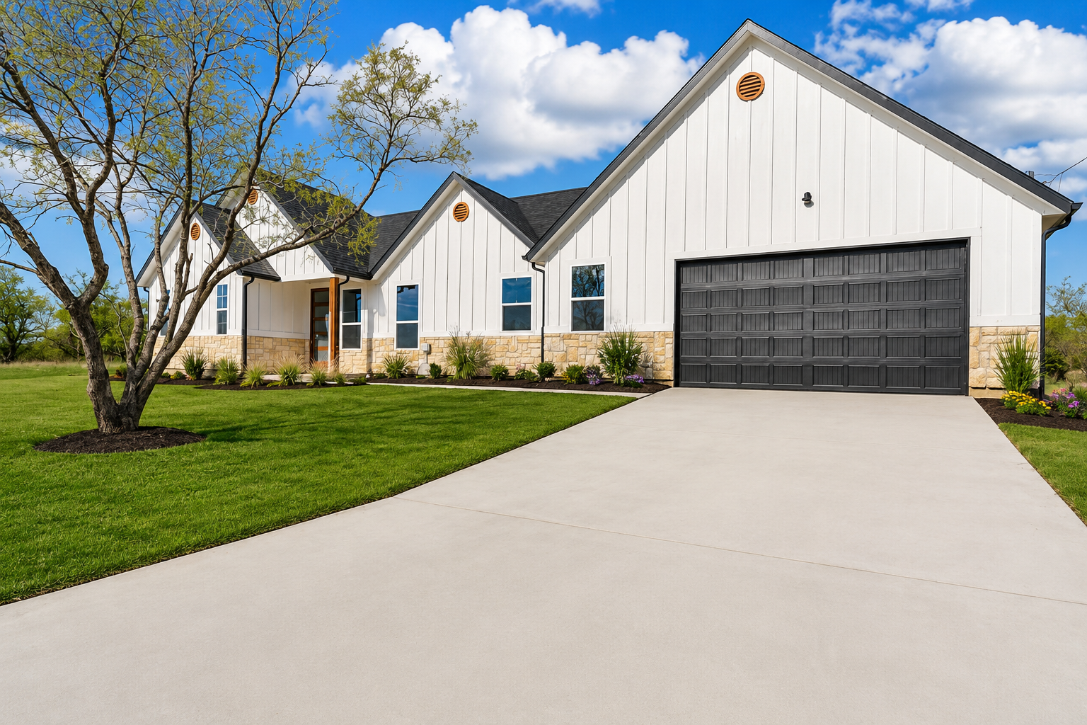 Modern house with white exterior and stone accents, surrounded by trees, with a concrete driveway leading up to a two-car garage on a clear day.