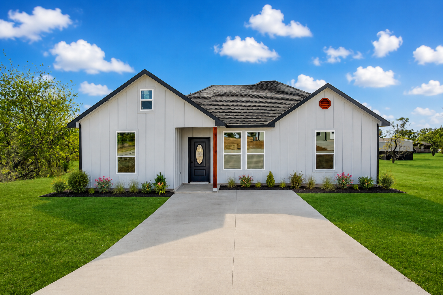A newly constructed white house with a black front door and a gray shingle roof, set against a bright blue sky with scattered clouds. The house is situated on a grassy lawn with a concrete driveway leading up to it.