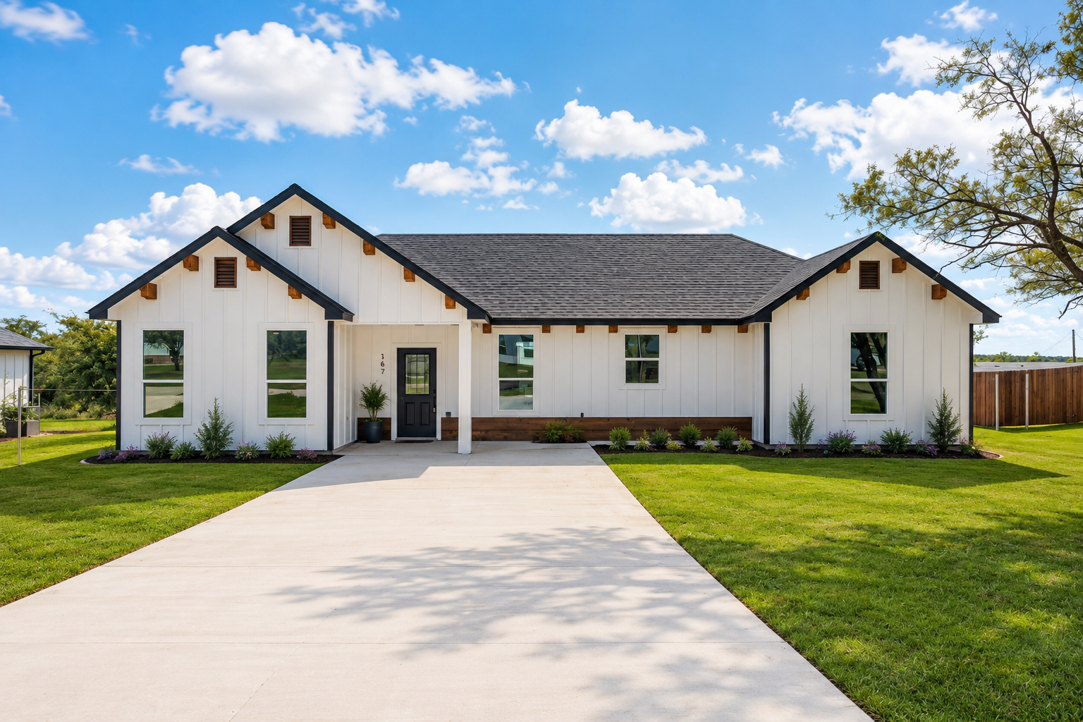 Newly constructed single-story house with a concrete driveway, dark gray and white exterior walls, and wooden window shutters. The house is still under construction, with dirt and building materials visible around it.