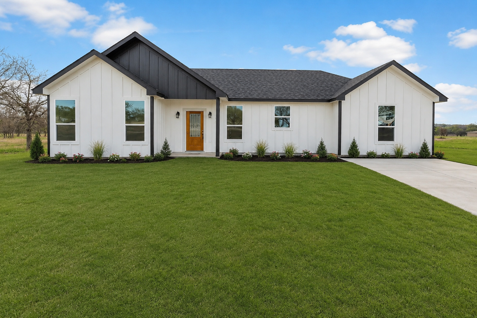 Newly built white house with black trim and a wood front door, surrounded by bare dirt yard and a concrete driveway on the right, in a rural area under a partly cloudy sky.