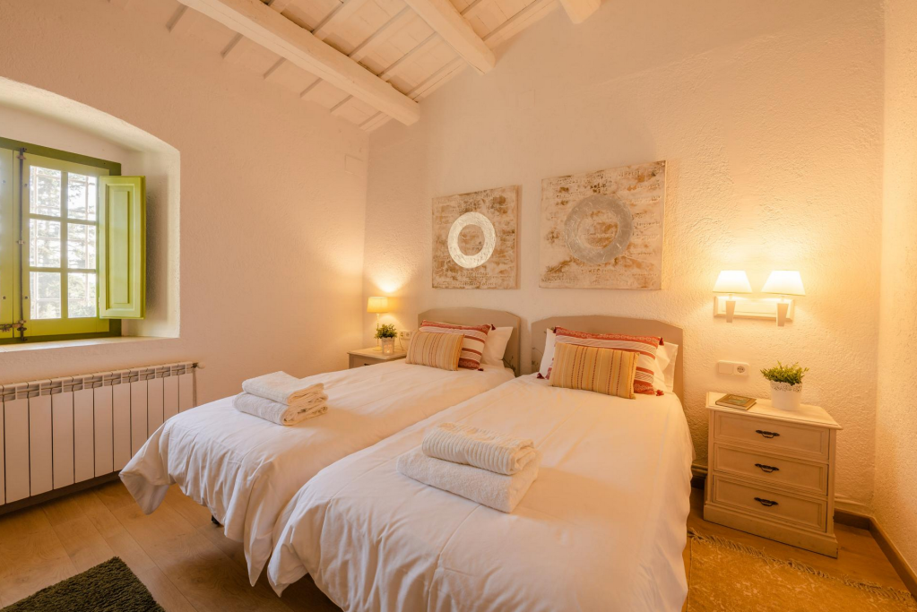 A cozy bedroom with two twin beds, each with white bedding and striped pillows. There are folded towels on each bed. The room has a small nightstand with a plant and a book, a wall-mounted light fixture, a window with green shutters, and artwork above the beds.