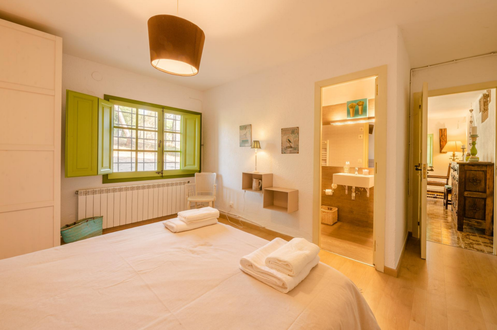 A bedroom with a large bed, green window shutters, a small chair, and a bathroom doorway. The room has wooden flooring and minimal decor.