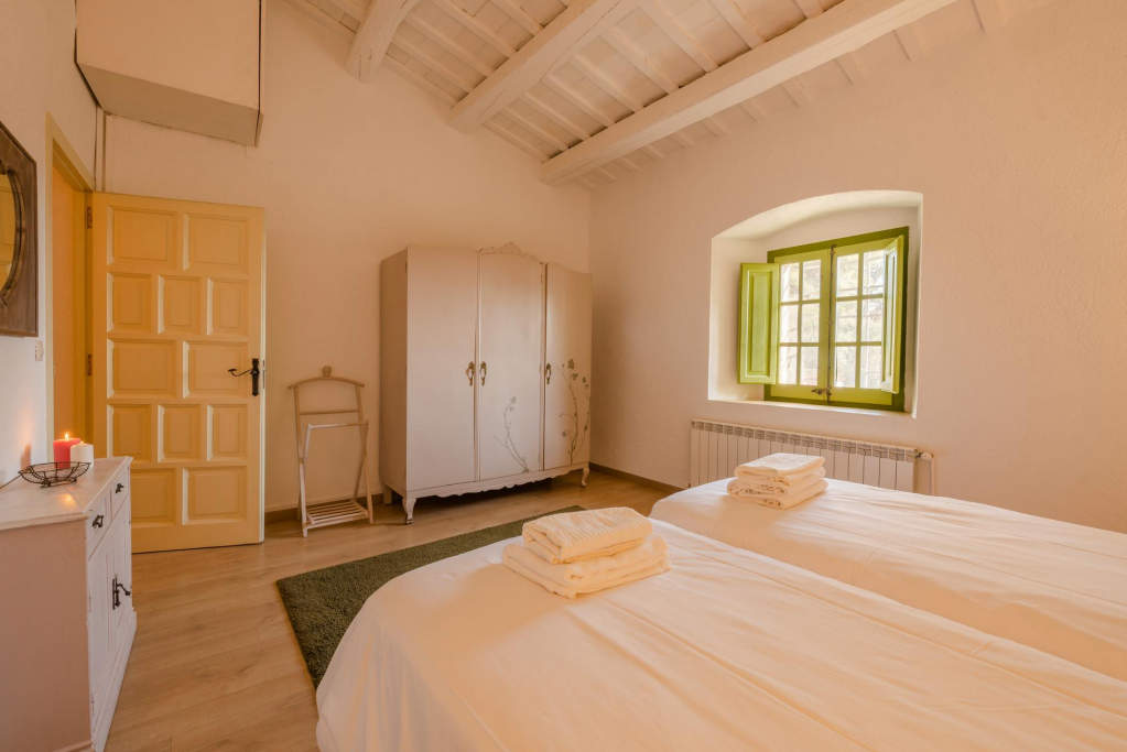 A cozy bedroom with two single beds covered in white linens and folded towels. There is a small white cabinet with candles, a white wardrobe, and a wooden folding chair. A green window with shutters lets in natural light, and the ceiling has white wooden beams.