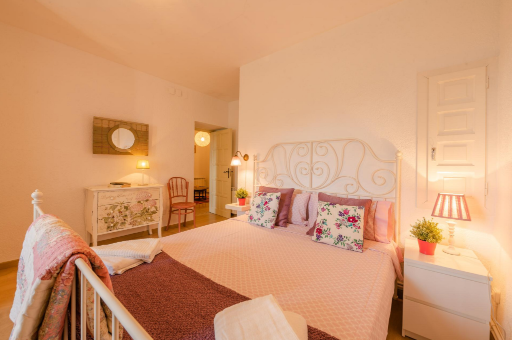 Cozy bedroom with a metal bed frame, pink bedding, floral pillows, white nightstand with a plant, and lamps; includes a small writing desk, chair, and decorative mirror.