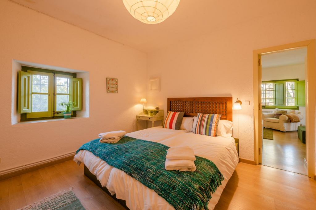 A cozy bedroom with a white bed, wooden headboard, multicolored striped pillows, green blanket, and folded towels. There is a small nightstand with a lamp, a window with green shutters, and a doorway leading to another room with a white sofa and matching green shutters.