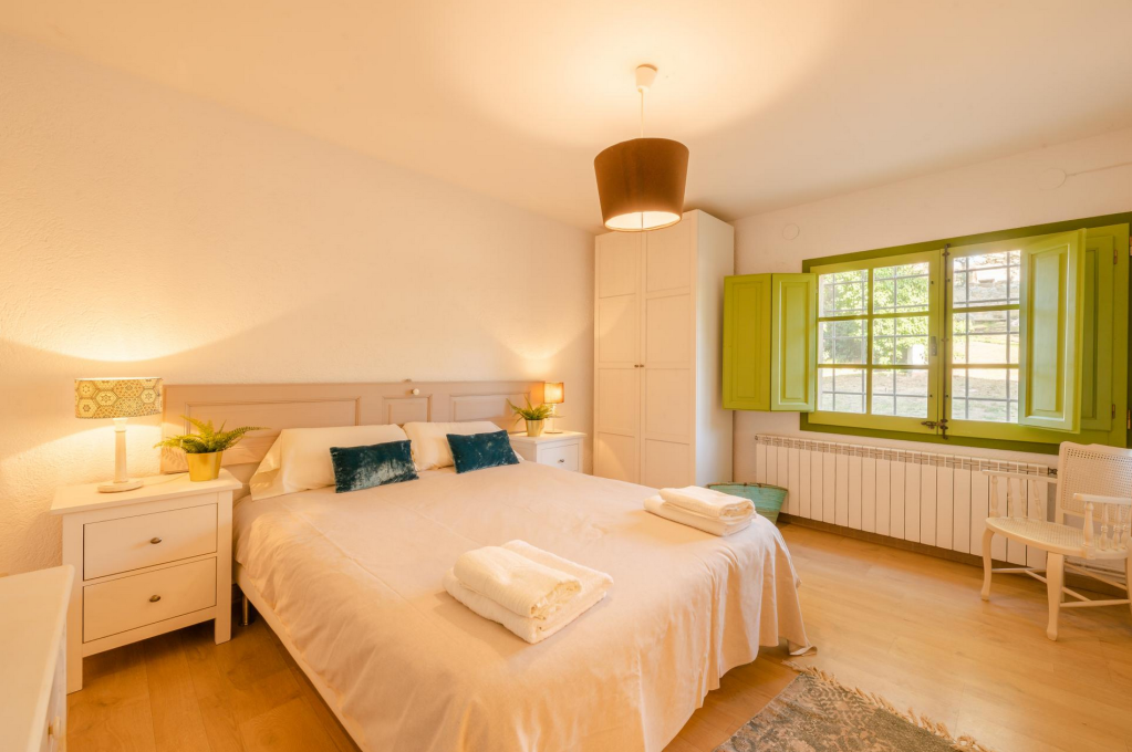 Bright bedroom with a large bed covered in beige linens, two folded towels on the bed, white nightstands with potted plants, two pillows, a white wardrobe, green window shutters, a white chair, and a hanging ceiling light.