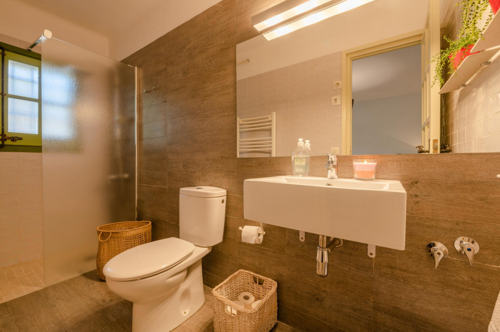Bathroom with frosted glass shower, toilet, small sink with toiletries, basket, towel heater, mirror, window, and plants.