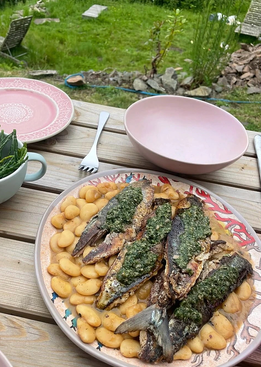 Plate of cooked fish topped with green herb sauce served over a bed of gnocchi on an outdoor wooden table, with empty bowls and utensils nearby.
