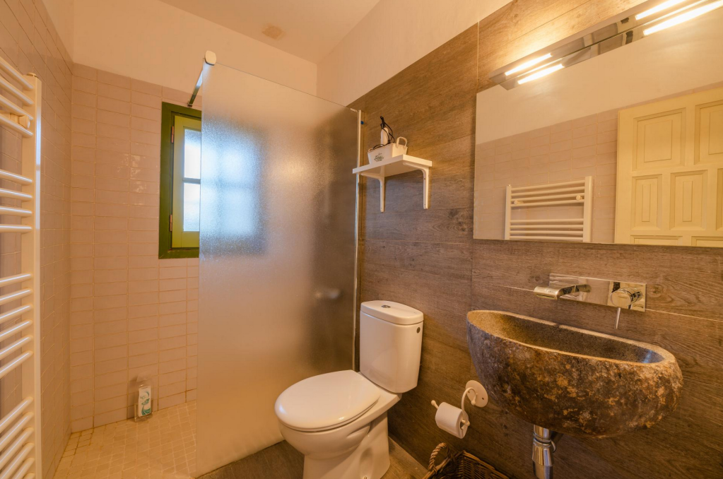 Bathroom with brown wooden wall tiles, a stone sink, a mirror, a toilet, and a frosted glass shower stall separated by a frosted panel. A small window, white shelf, and towel radiator are also visible.