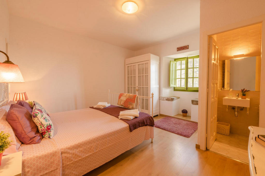 A cozy bedroom with a bed covered in pink bedding, floral pillows, and a small side table with a lamp. There is a white wardrobe, a window with green shutters, and a small area with a rug and built-in seats. A doorway leads to a bathroom with a sink and mirror.