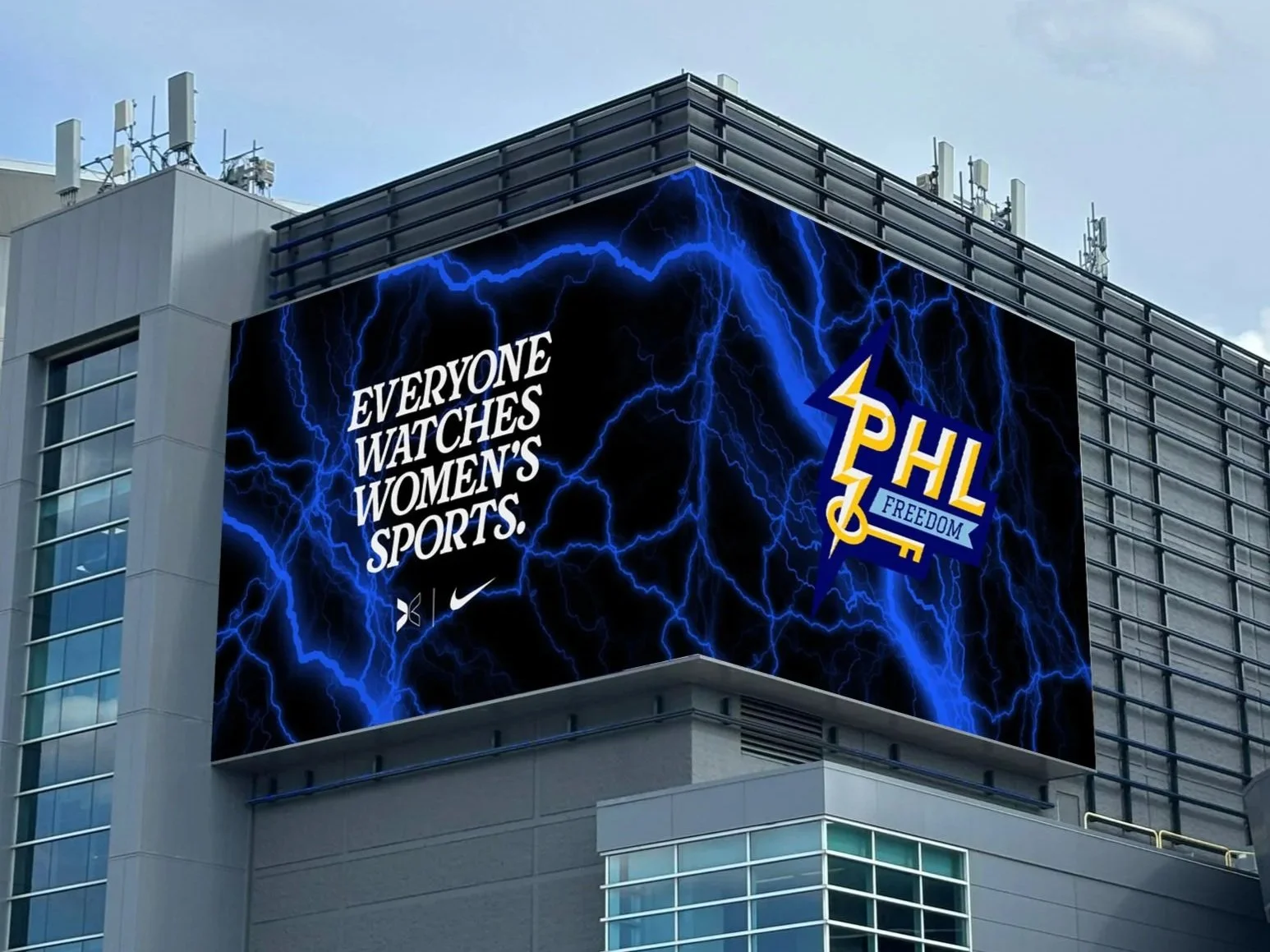 Large digital billboard on the side of a modern building reading “Everyone watches women’s sports” beside the PHL Freedom lightning logo, with blue lightning graphics across a black background.