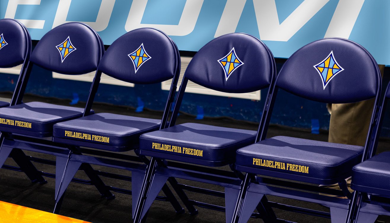 Row of navy courtside folding chairs branded “Philadelphia Freedom,” each with the team’s diamond logo on the backrest, lined up along the sideline of a basketball court.