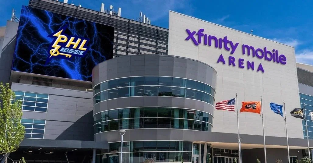 Exterior of Xfinity Mobile Arena with a large digital billboard displaying the PHL Freedom lightning logo, under a clear blue sky with flags flying in front of the building.