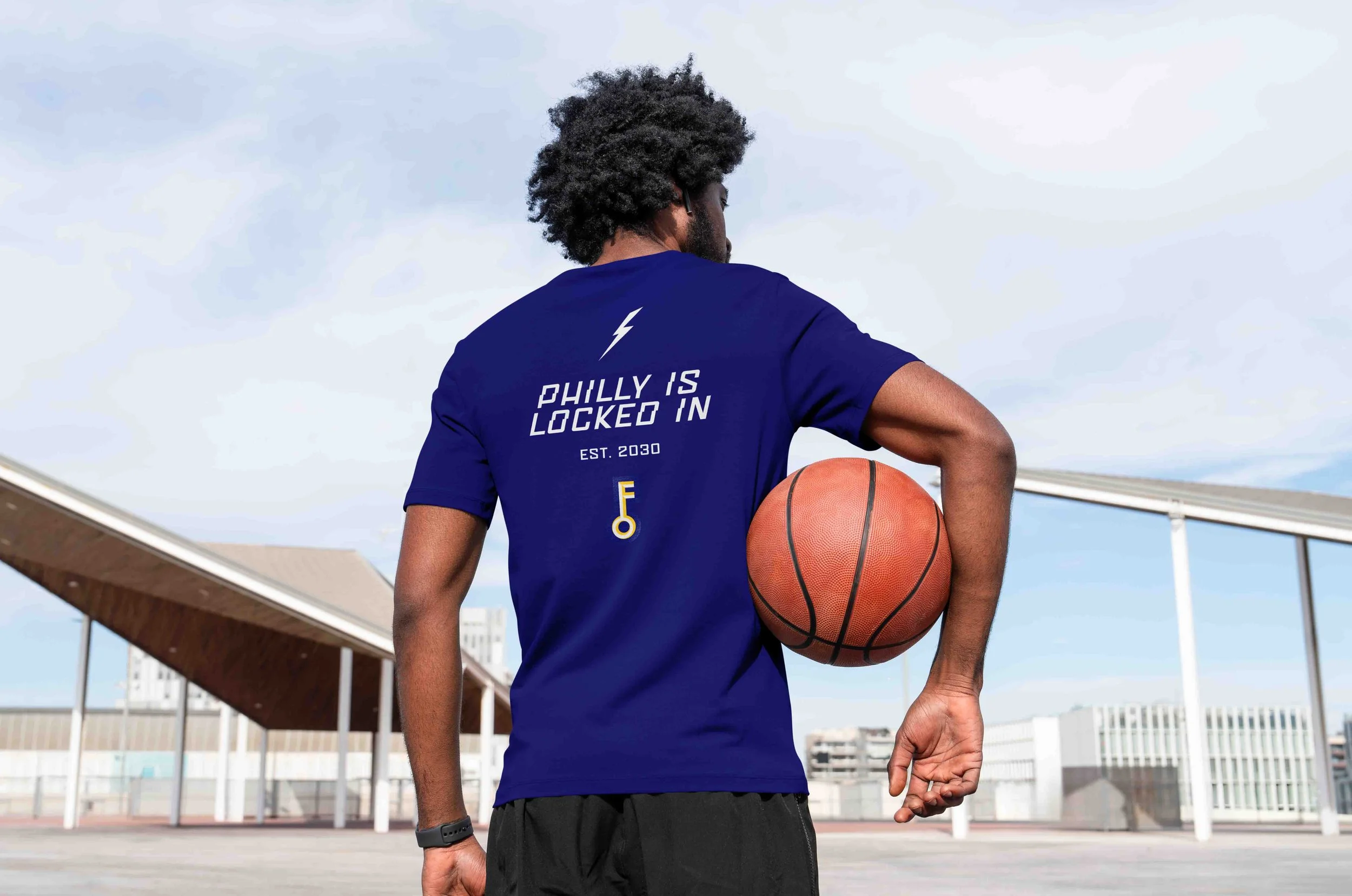 Back of man holding basketball under his arm in a dark blue T-shirt featuring white custom lettering reading “Philly is locked in, est. 2030” with a yellow “F” key logo beneath, photographed in an urban outdoor setting.