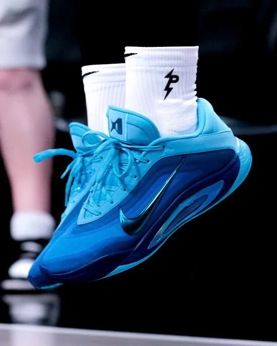 Close-up of a player’s feet on a basketball court wearing blue A’One basketball shoes and white crew socks with “P” lightning logo, with the hardwood floor visible beneath.