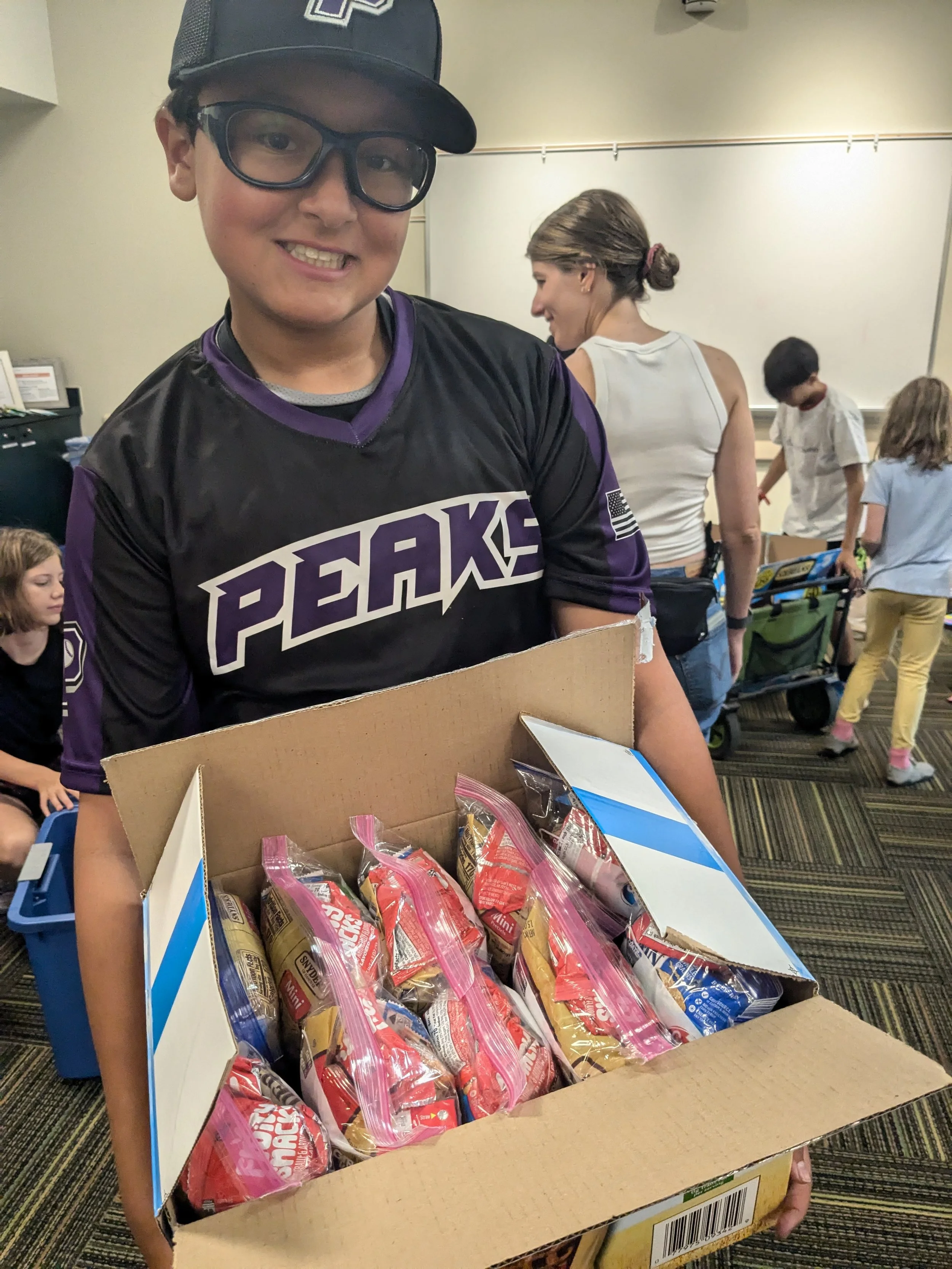 A boy wearing glasses, a black and purple PEAKS sports jersey, and a cap, holding an open cardboard box filled with packaged snacks, in a room with other children and adults.