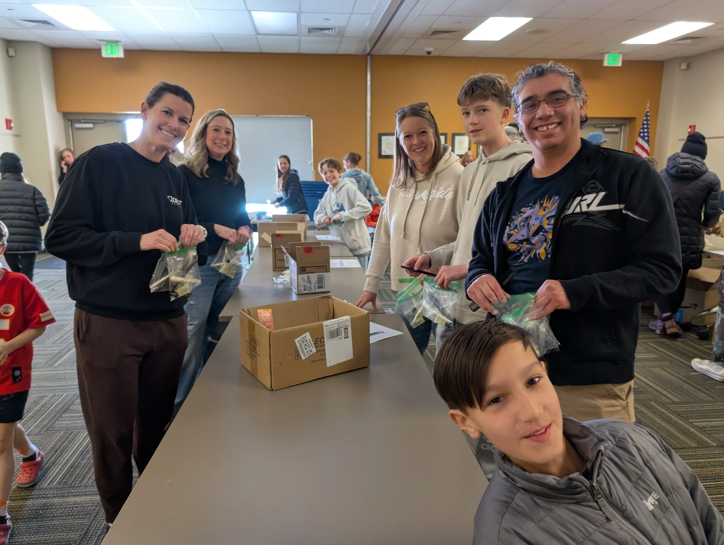 Group of volunteers packing food in plastic bags at a community event in a room with yellow walls and people in the background.