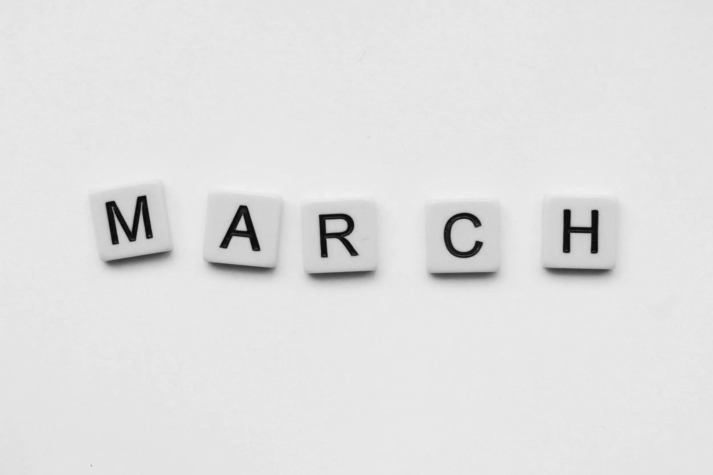 Black and white photo of letter tiles spelling 'MARCH' on a plain white background.