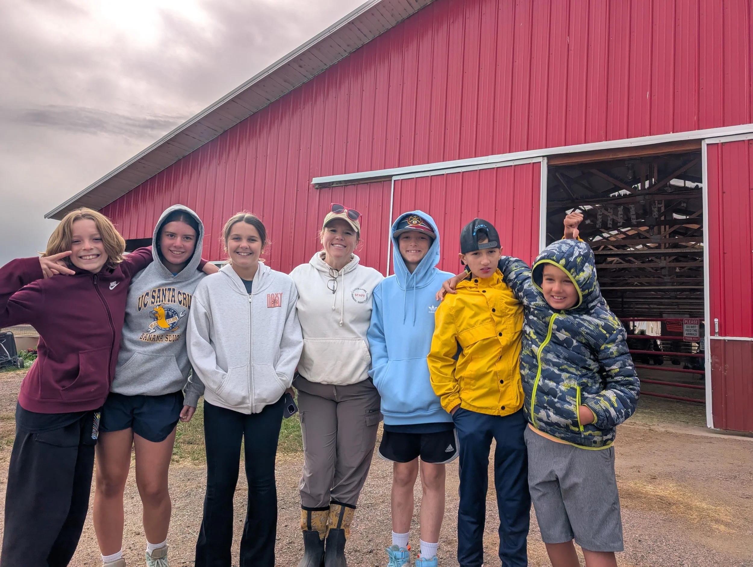 Group of seven children standing in front of a large red barn with clouded sky above, smiling and posing for the photo.