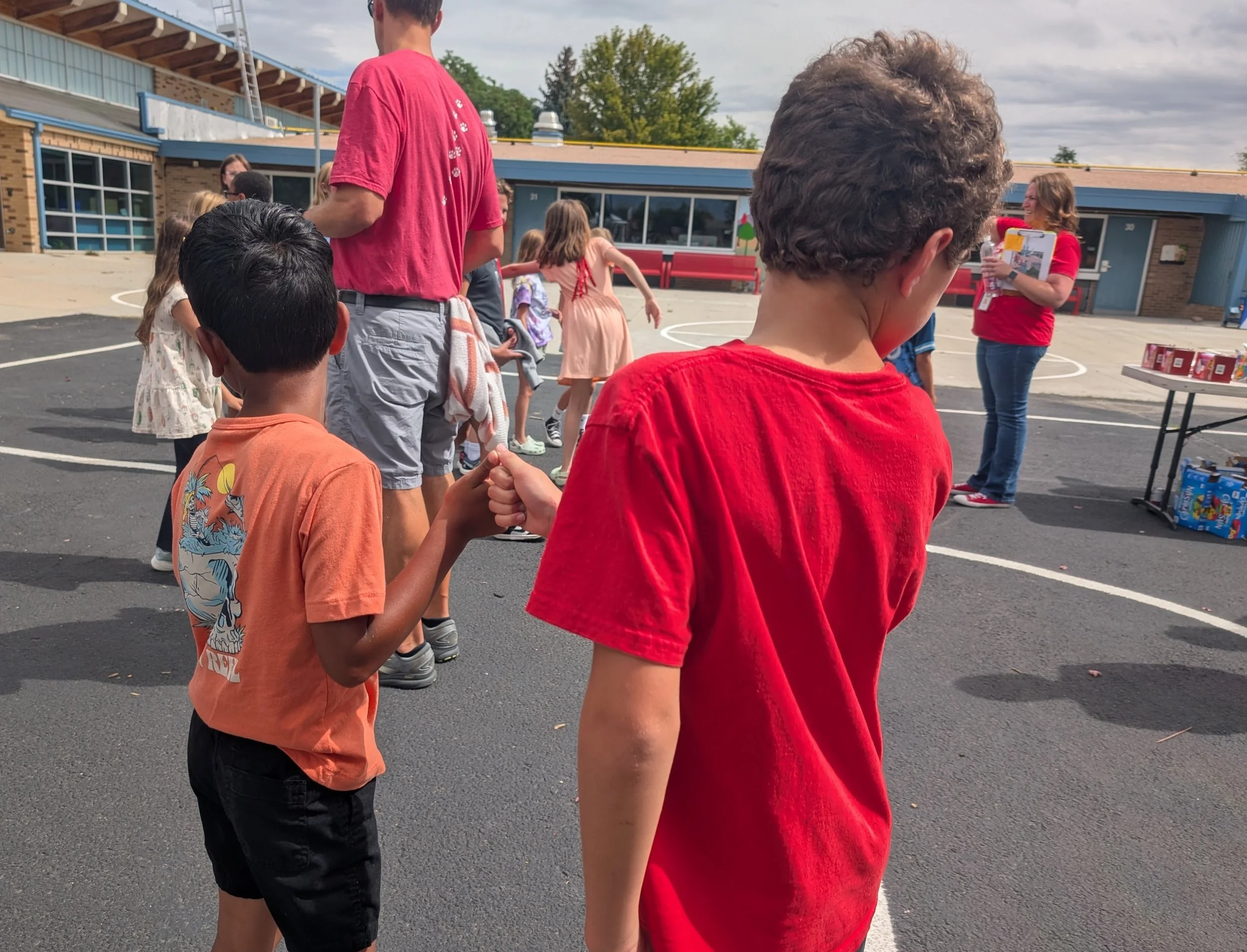 Children and adults gathered outdoors near a school building, participating in a fist bump or handshake line, with a table of snacks or prizes on the right side.