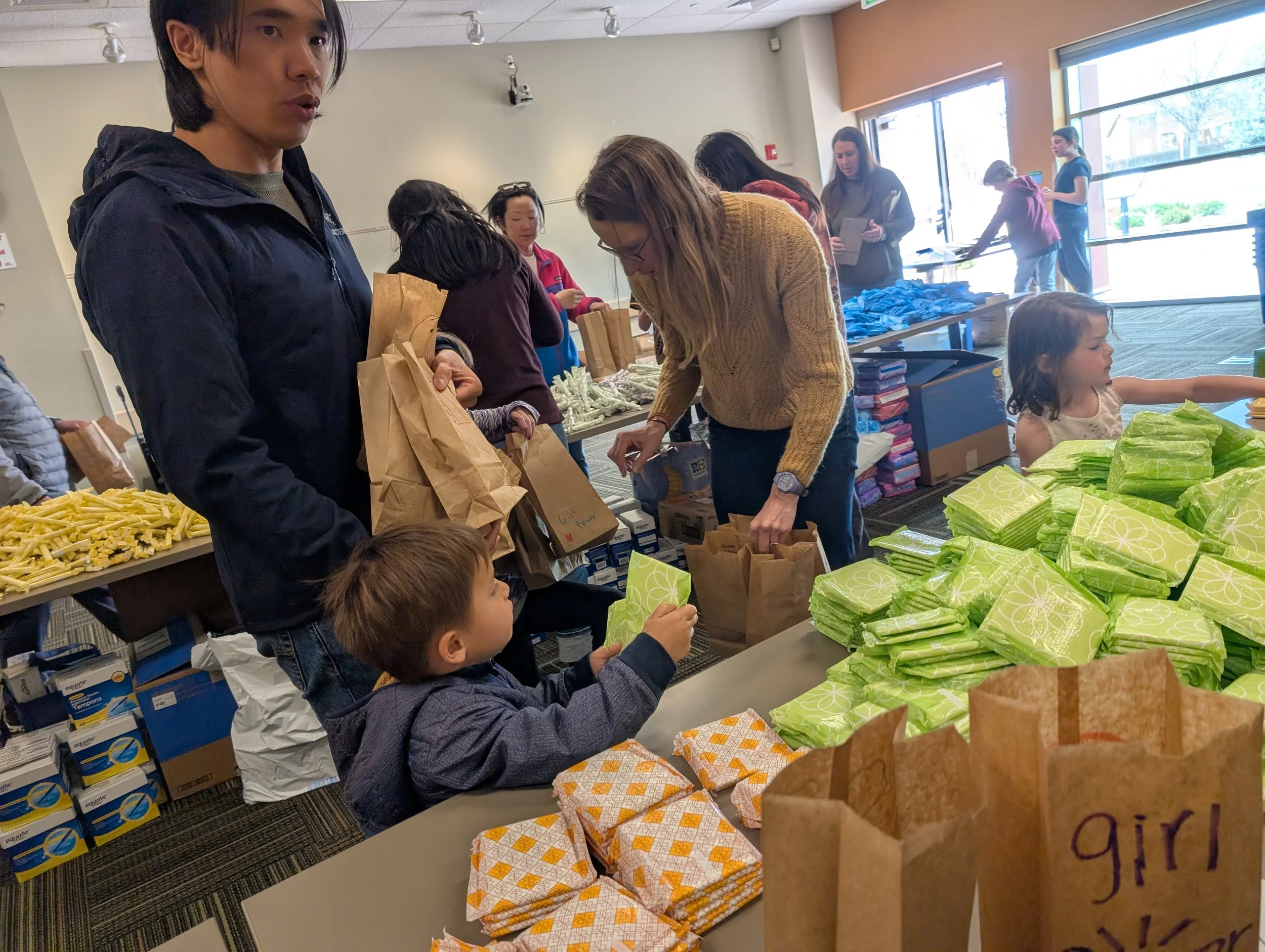 People volunteering at a charity event, filling bags with snack items. Children and adults are working together in a well-lit room, with tables filled with wrapped snacks and supplies.