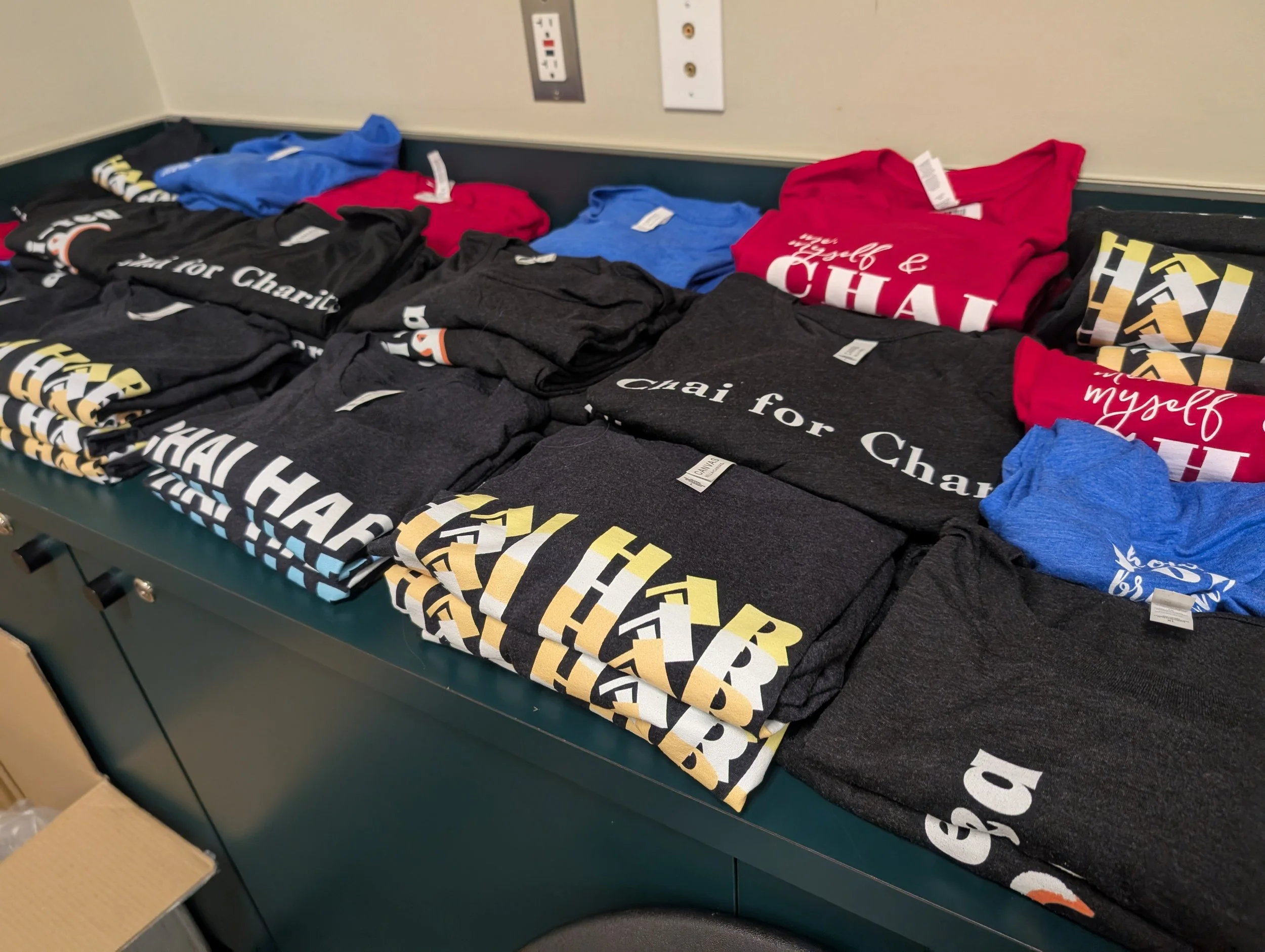 A table displaying folded T-shirts with various slogans and colors, including black, gray, blue, and red, in a retail or donation setting.