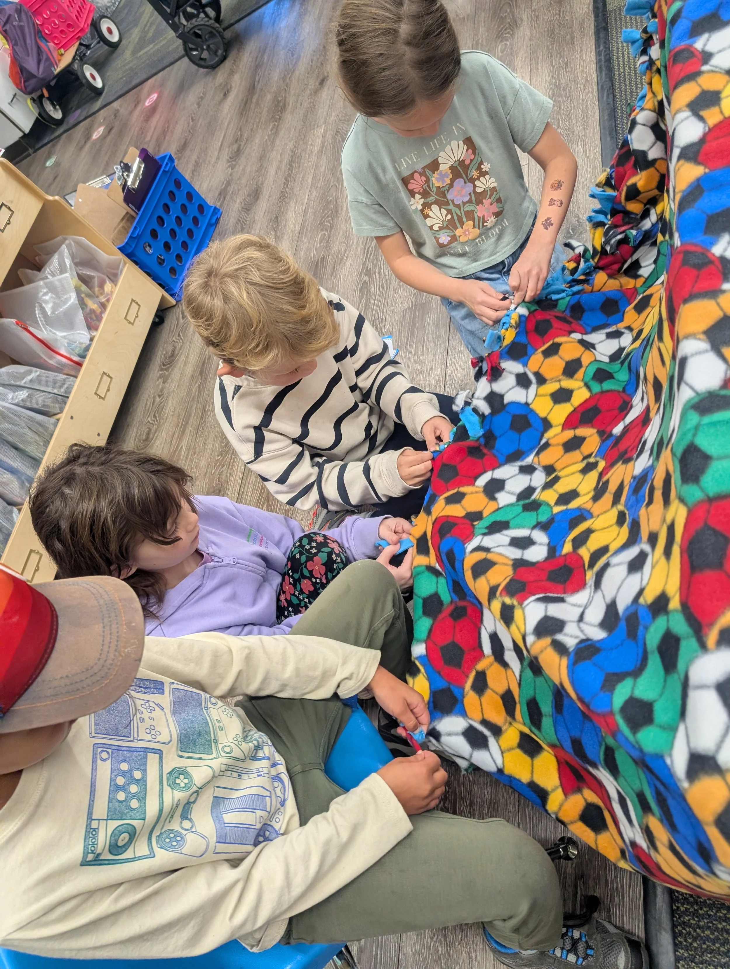 Children working on a colorful blanket with soccer ball patterns inside a store or workshop.