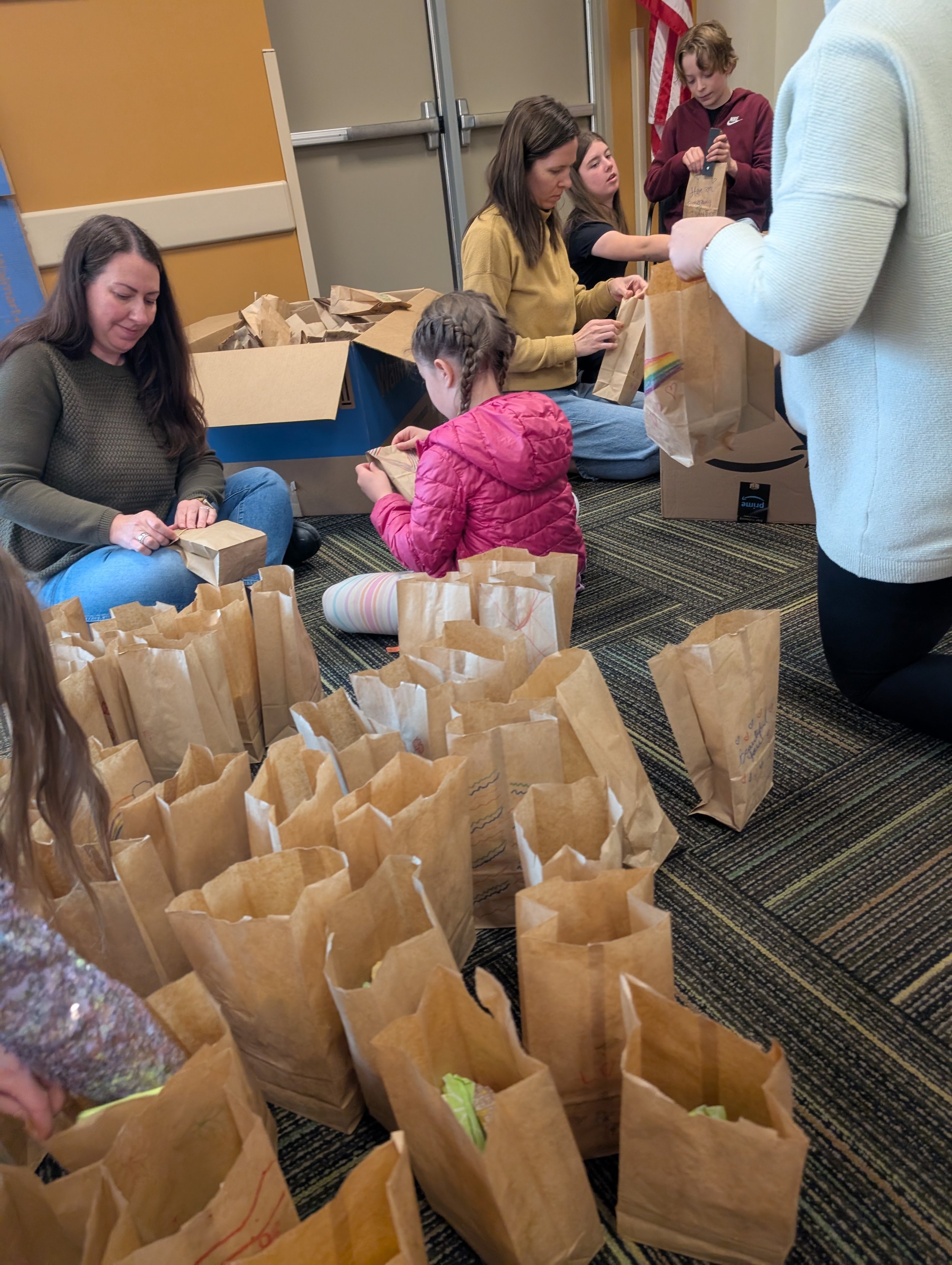 Group of people, including children and adults, sitting and kneeling on the floor, packing brown paper bags, with a large box of additional paper bags nearby, in an indoor setting with beige and orange walls and an American flag in the background.