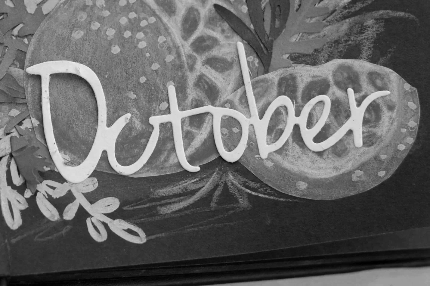 Decorative sign with the word 'October' written in cursive, placed on a dark surface with leaves and floral drawings surrounding it.
