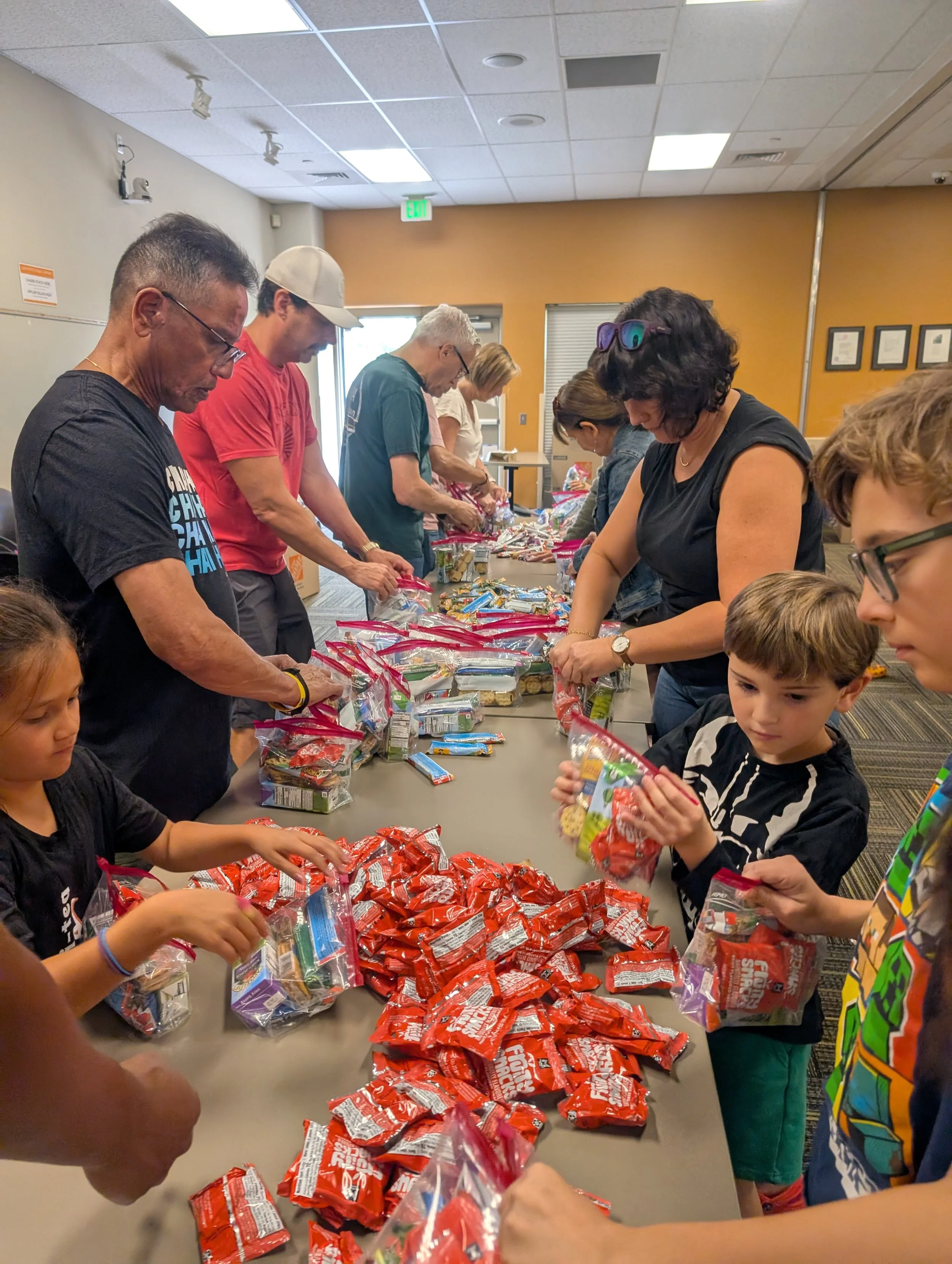 People, including children and adults, gather around a table sorting and packing various snack bags in a room with orange walls.