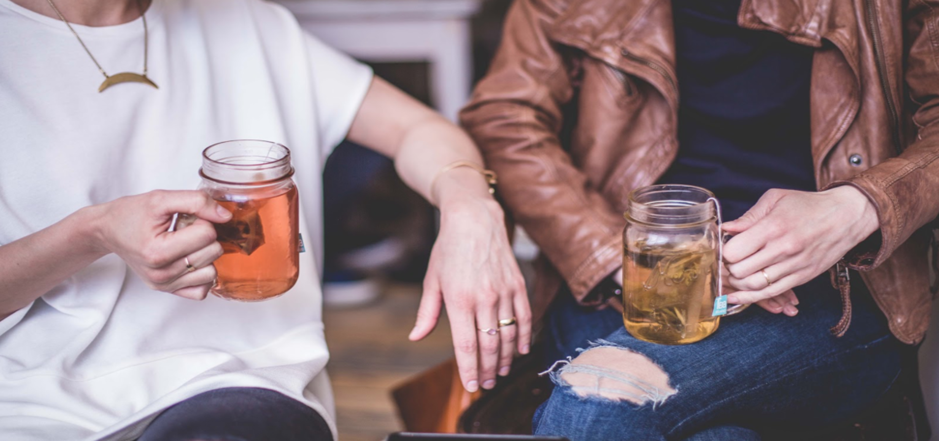 Two people sitting together, each holding a jar mug of tea or herbal drink, with one person wearing a white shirt and the other in a brown leather jacket.