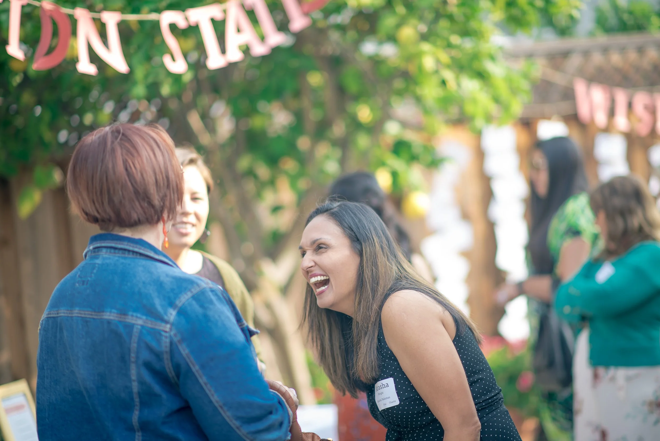 Two women laughing and talking at an outdoor event with other people in the background, greenery, and a sign that partly says 'IN STALL'.