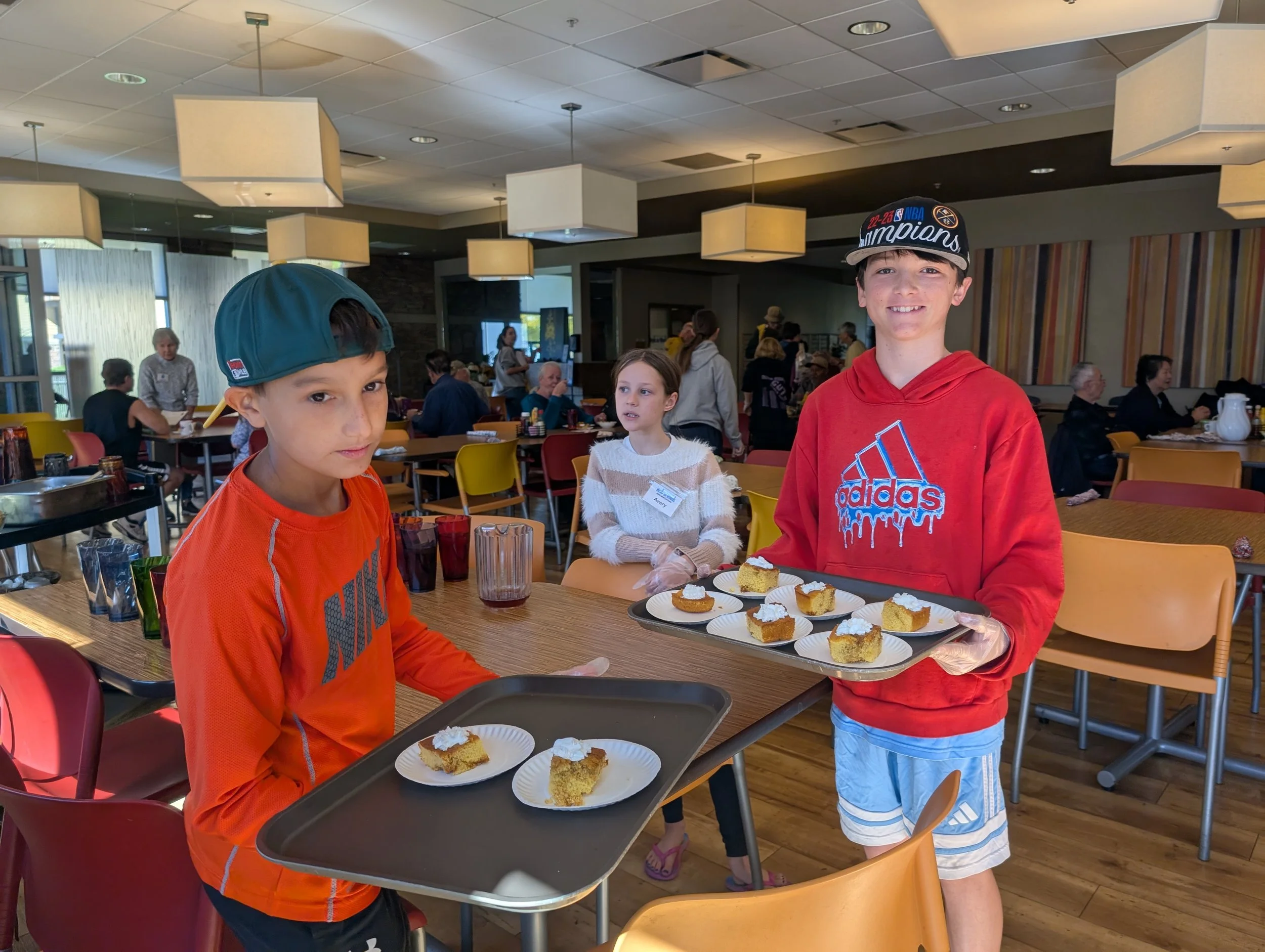 Two young boys holding trays with pieces of cake in a restaurant or cafe, with a girl in the background and other patrons at tables.