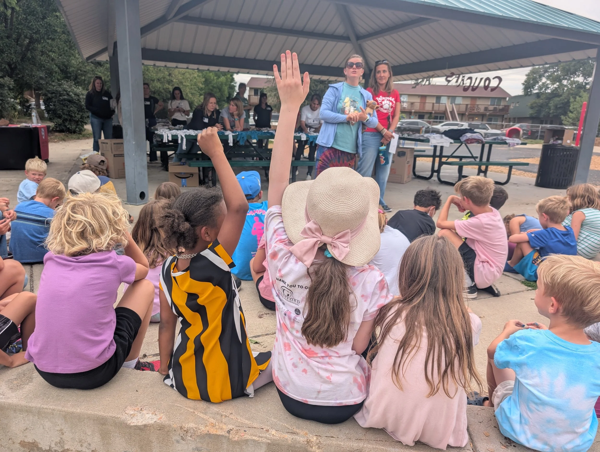 Children sitting on a concrete ledge outdoors, engaging in a presentation or performance under a pavilion, with some raising their hands to ask questions, while adults stand in the background near tables with merchandise.