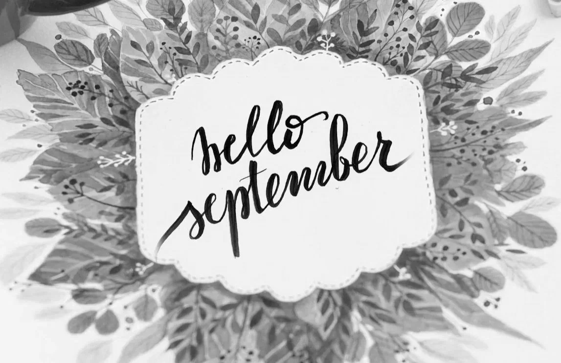 A decorative black and white illustration with the words 'hello September' in cursive font centered inside a scalloped shape, surrounded by leafy foliage.