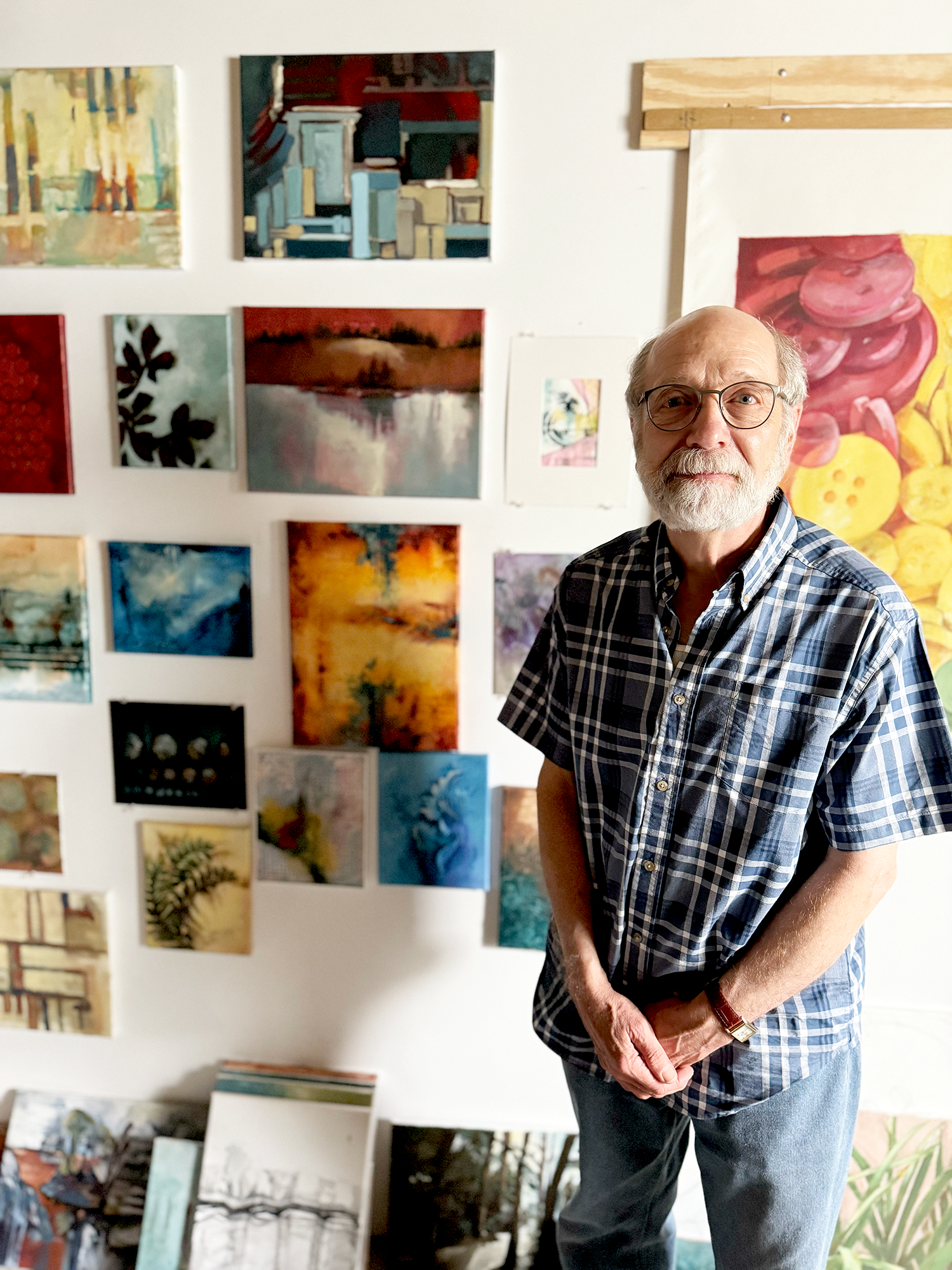 An elderly man with glasses and a checkered shirt standing in front of a wall filled with small, colorful paintings, and some artwork on the floor.