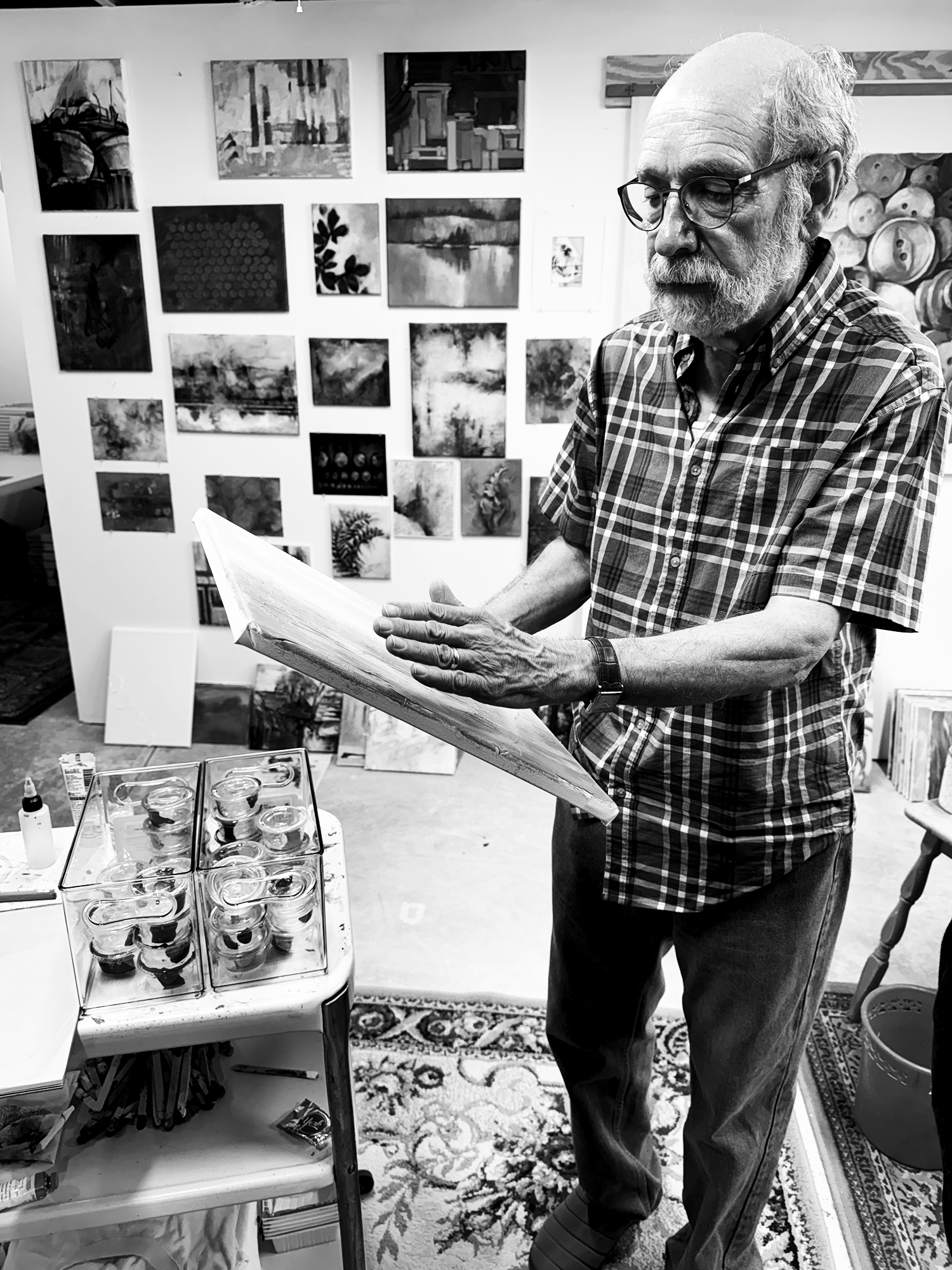 An elderly man with glasses and a beard, wearing a checkered shirt, is examining a canvas in a studio. Behind him is a wall decorated with various artworks, and in front of him, a small table holds several jars and art supplies.