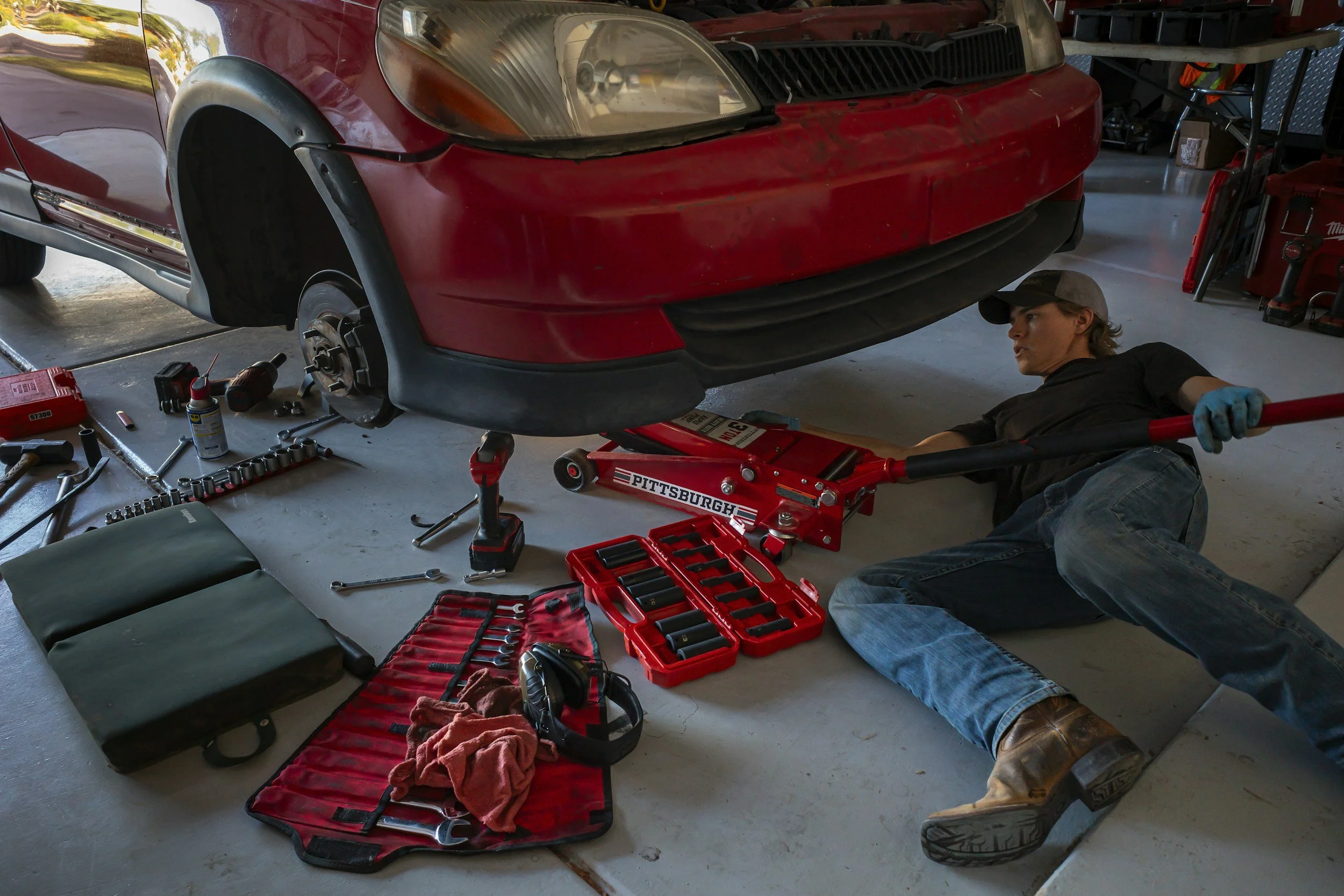 A mechanic working on a red car lifted with a floor jack in a garage. The mechanic is lying on the floor, wearing a baseball cap, black shirt, jeans, and brown boots, using a long tool. Various tools and equipment are scattered around, including wrenches, a red tool kit, a green seat cushion, and a headlamp.