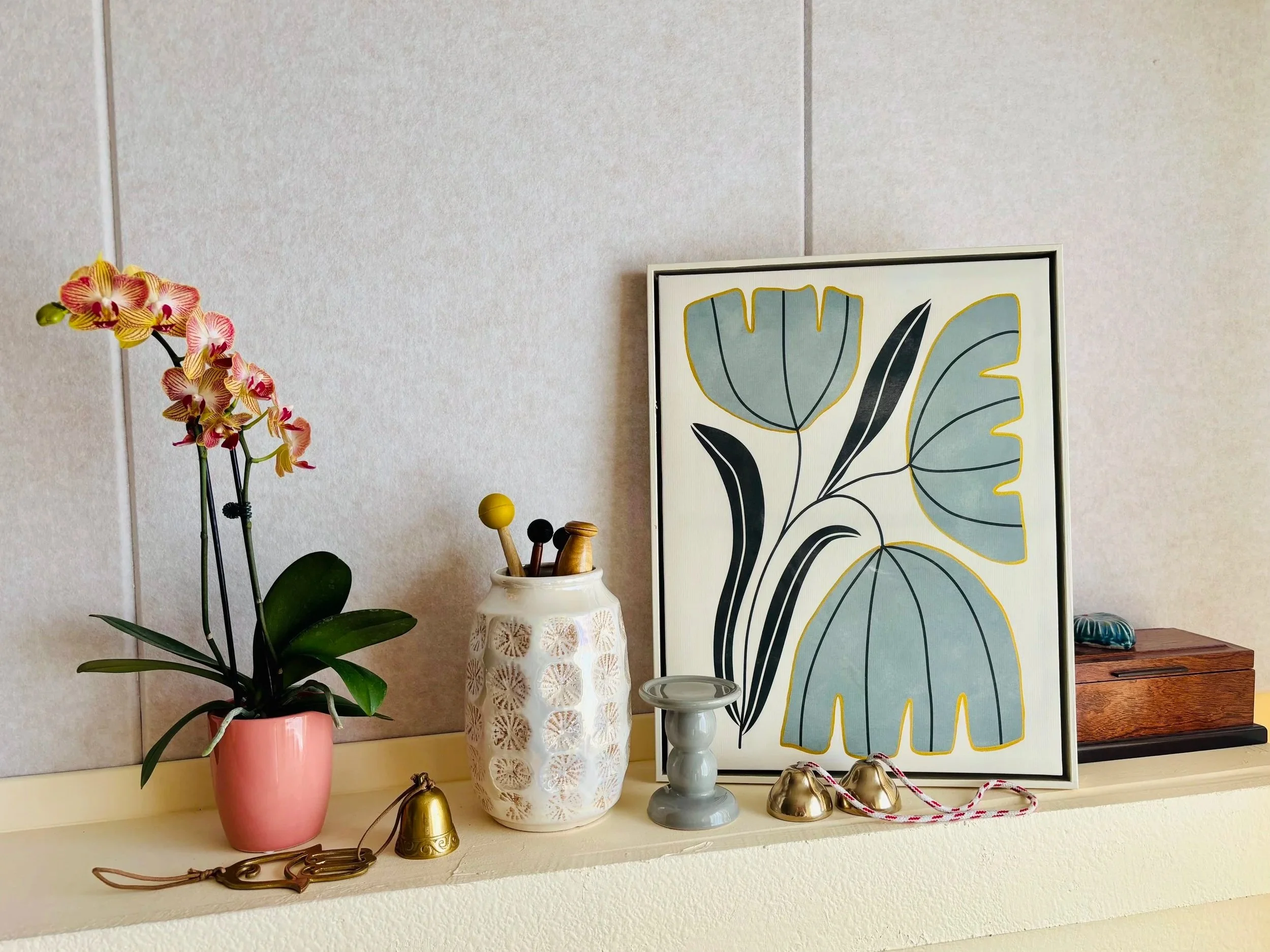 A decorated shelf with an orchid plant in a pink pot, a white vase holding small decorative objects, a framed botanical artwork, a small gray pedestal, gold bells, a wooden box, and a shell. kindred yoga mykindred yoga kindrd yogaa my kindred yoga