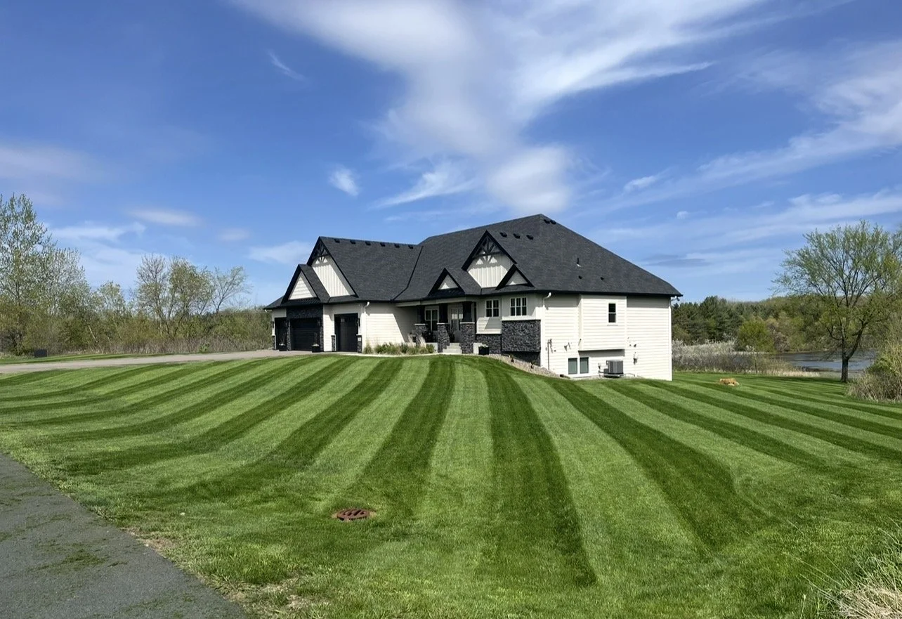 A modern house with black roof and white siding, surrounded by neatly mowed green grass with striped pattern, and trees in the background under a partly cloudy blue sky, freshly mowed lawn managed by Ornell lawn services. 