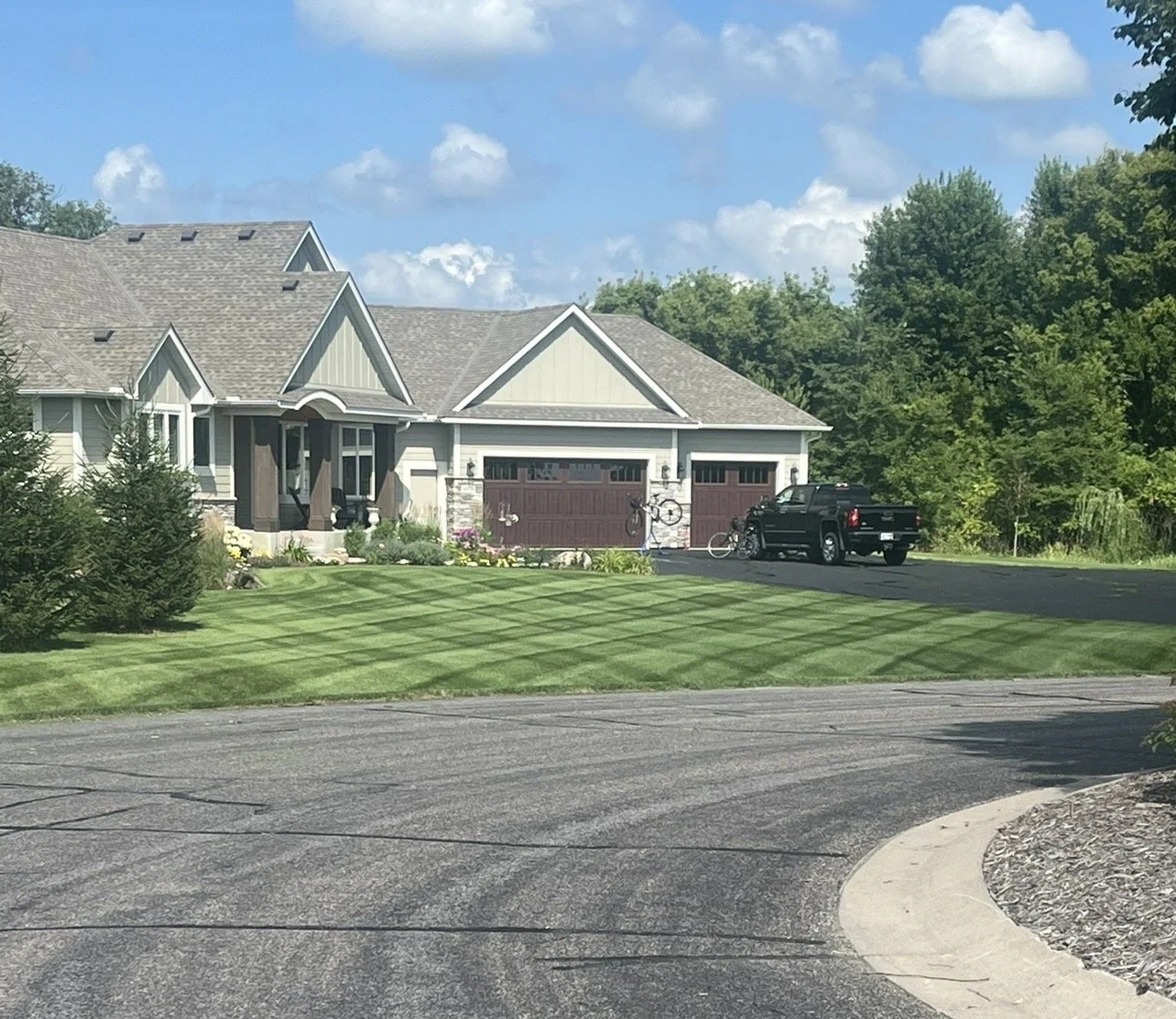 A suburban house with a well-maintained lawn by Ornell Lawn Services, a driveway with a parked pickup truck, bicycles, and surrounded by green trees under a partly cloudy sky.