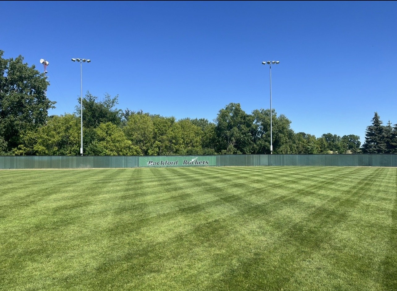 A baseball or softball field with a well-maintained grassy outfield managed by Ornell Lawn Services, a green outfield fence with a sign that reads 'Rockford Rockets', and tall floodlights under a clear blue sky with trees in the background.
