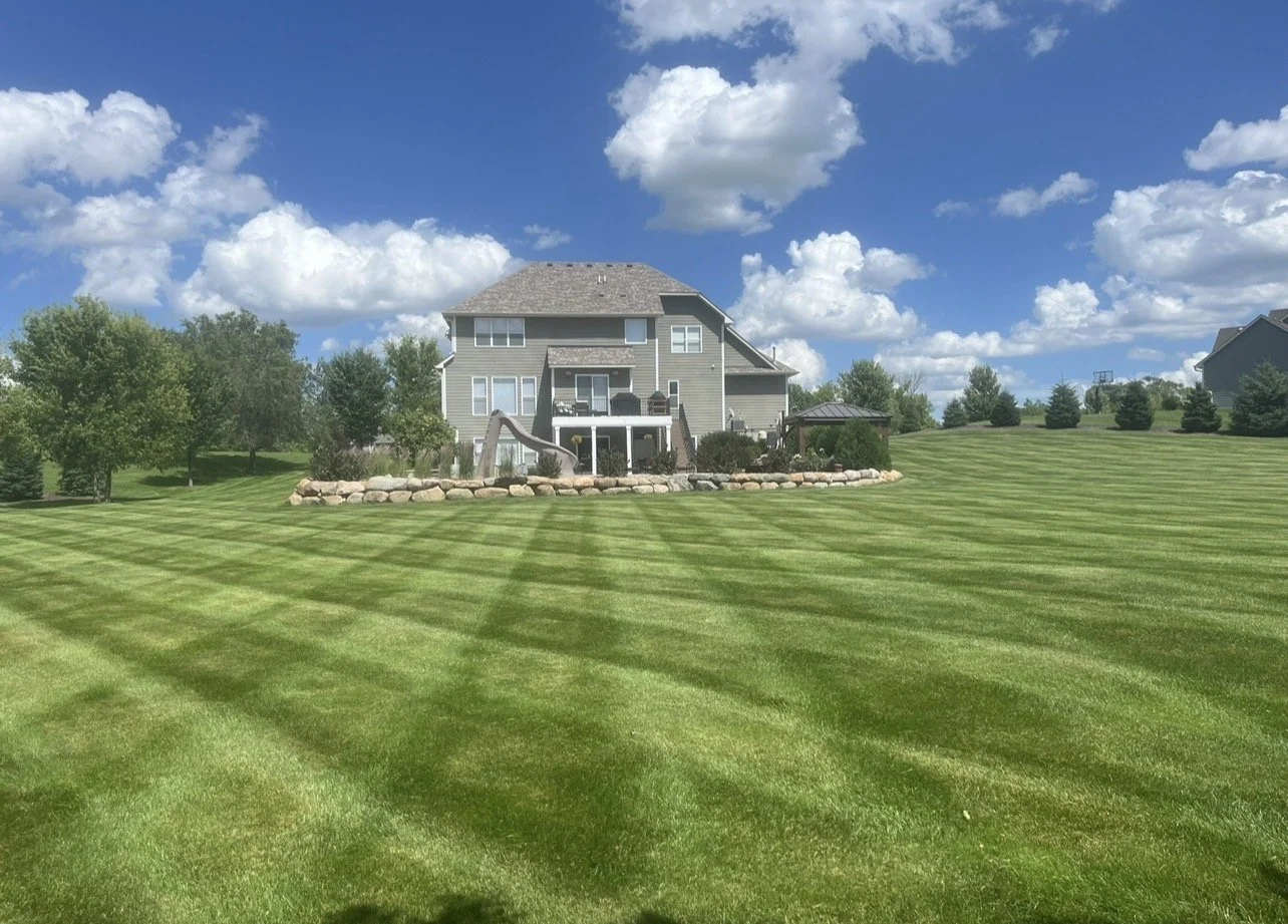 A large backyard with neatly mowed grass by Ornell Lawn Services, trees on the perimeter, and a gray house with a balcony and a slide leading into the yard.