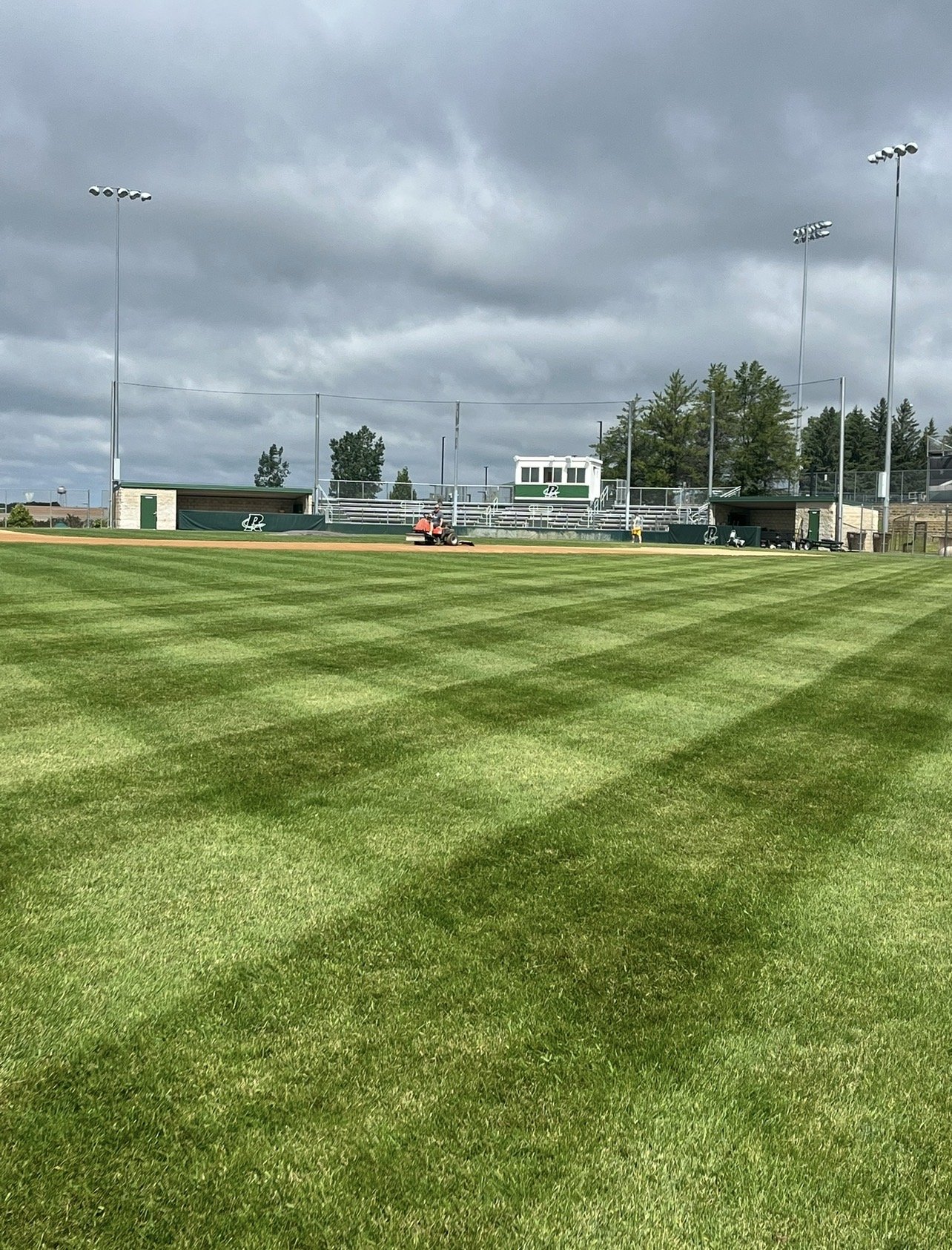 A baseball field with well-maintained green grass by Ornell Lawn Services, stadium lights, and a cloudy sky in the background. A person is mowing the field.