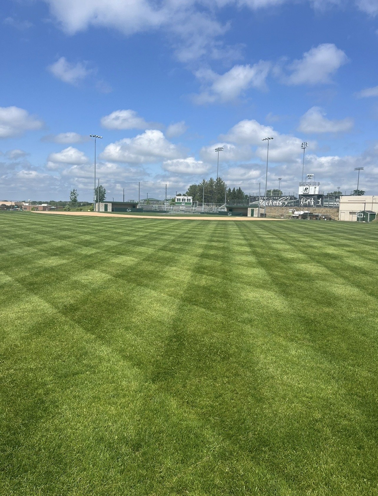 Empty baseball field under a blue sky with white clouds, with green grass and stadium lights in the background. Freshly mowed lawn managed by Ornell lawn services. 
