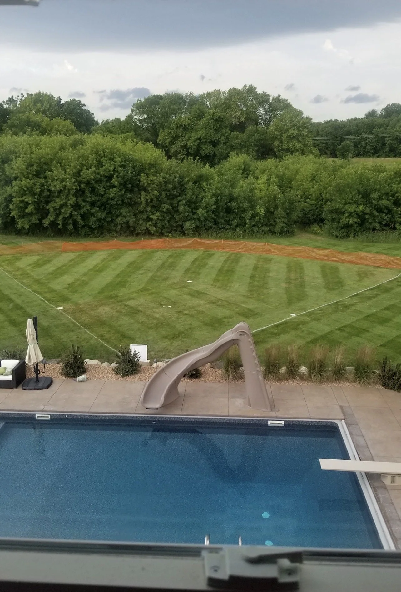 A backyard view through a window showing a swimming pool, a slide, a patio with outdoor furniture, a grassy field with striped mowing patterns freshly mowed by Ornell Lawn Services, construction fencing, and a background of trees and a cloudy sky