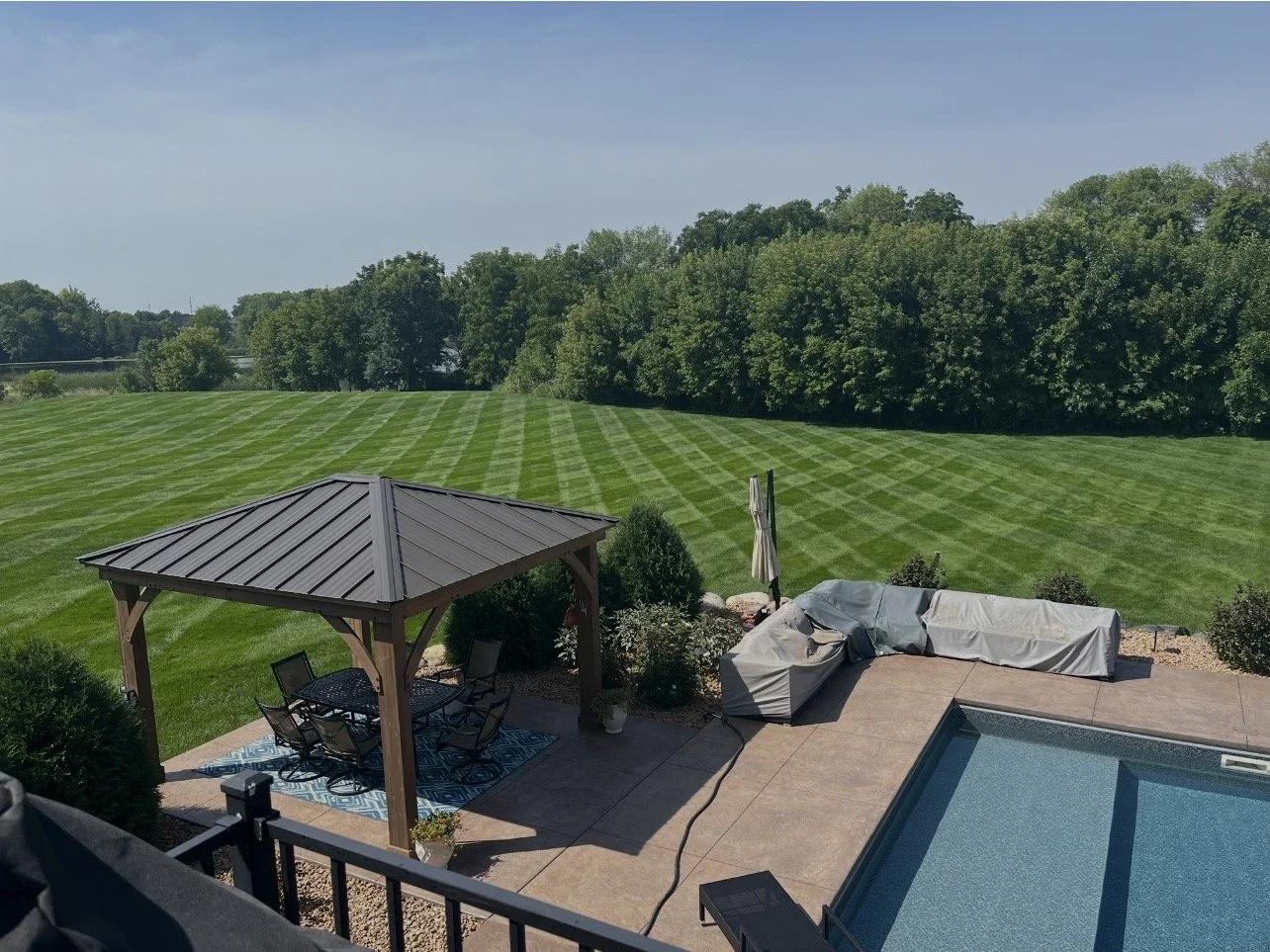Backyard with a swimming pool, patio furniture under a gazebo, and an open green field with trees in the background and freshly mowed lawn by Ornell lawn services. 